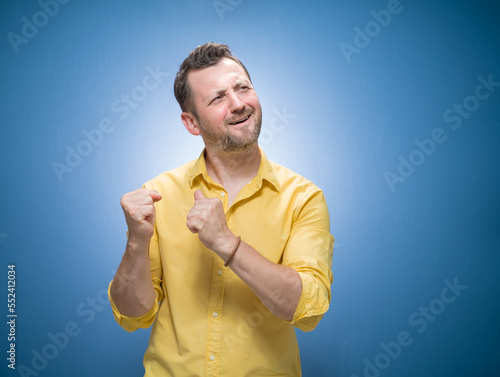 Amused handsome young man with closed eyes and clenched fists over blue background, dresses in yellow shirt. Yes concept. Amusing guy celebrates success