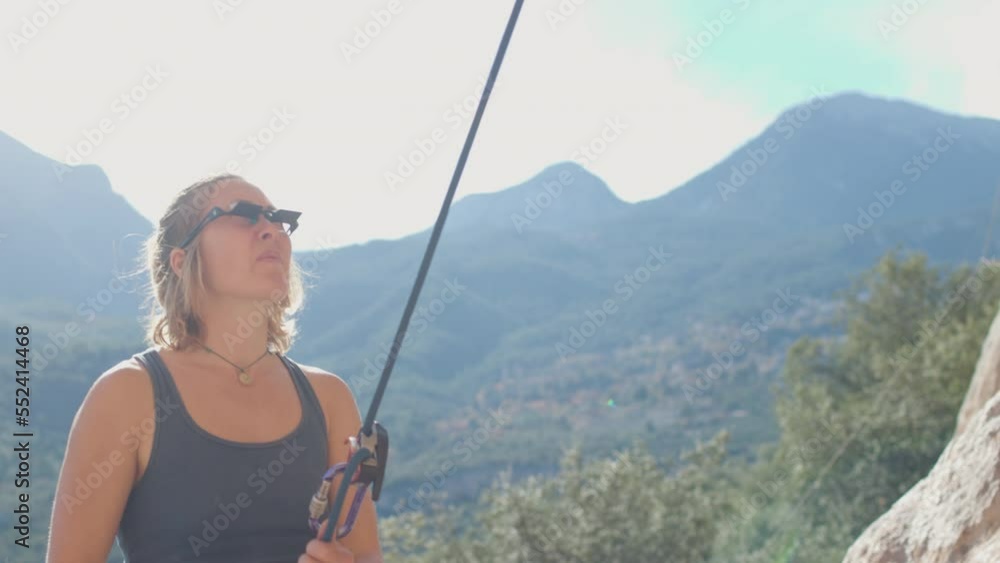 A rock climber stands in a canyon, holding a rope. the climber insures his partner. Sports girl smiles and holds climbing equipment. Climber near the cliff. A female climber is preparing to climb the A rock climber stands in a canyon, holding a rope. the climber insures his partner. Sports girl smiles and holds climbing equipment. Climber near the cliff. A female climber is preparing to climb the