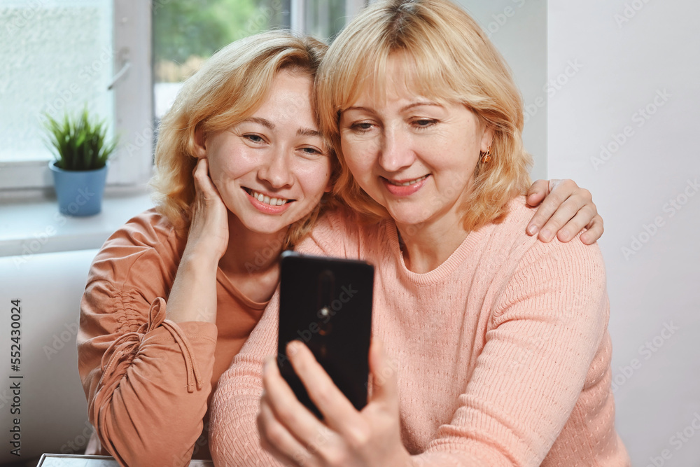 Happy adult daughter and mother taking a selfie or using smartphone and making video call, smiling at the camera. Home interior, natural appearance. Mother and daughter spending time together. Happy adult daughter and mother taking a selfie or using smartphone and making video call, smiling at the camera. Home interior, natural appearance. Mother and daughter spending time together.