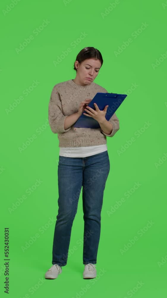 Vertical video: Front view of casual person looking at clipboard papers on full body greenscreen, taking notes on files and working on analysis. Female model writing information on documents in studio Vertical video: Front view of casual person looking at clipboard papers on full body greenscreen, taking notes on files and working on analysis. Female model writing information on documents in studio
