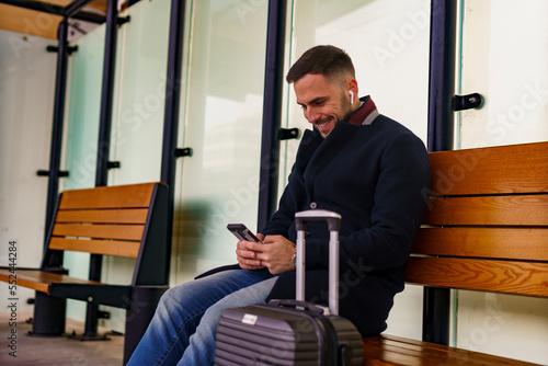 Young man on a train station smiling at his phone, sitting next his suitcase, he's going on a business trip