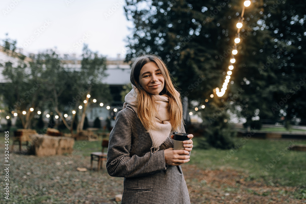 Young beautiful girl holding a cup of coffee in her hands on the street in the park with lights in a warm coat with a scarf Young beautiful girl holding a cup of coffee in her hands on the street in the park with lights in a warm coat with a scarf