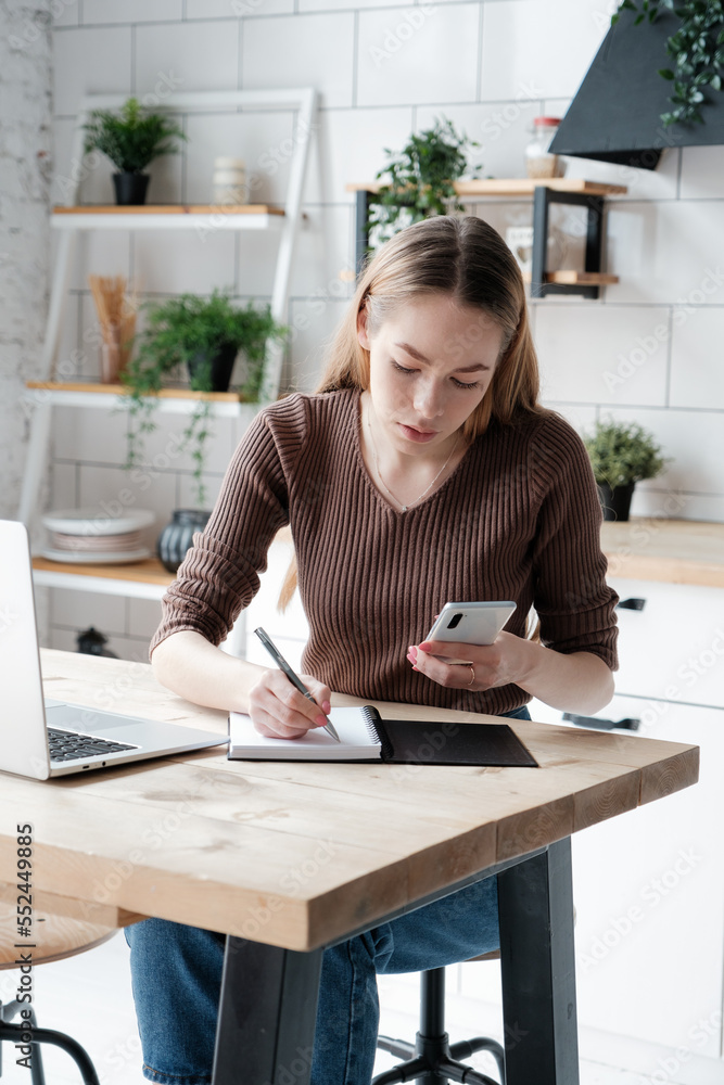 Young caucasian woman looking on smartphone and writing in notepad while sitting at the desk and working on remote project at home Young caucasian woman looking on smartphone and writing in notepad while sitting at the desk and working on remote project at home