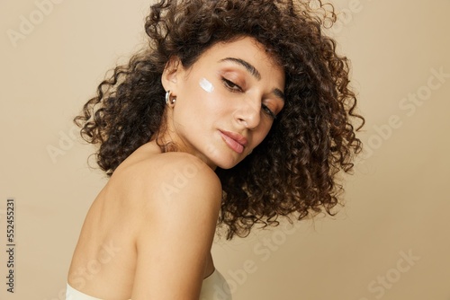 Woman beauty face close-up applying anti-aging moisturizer with fingers of her hand, skin health nails and hair, hair dryer style curly afro hair, body and beauty care concept