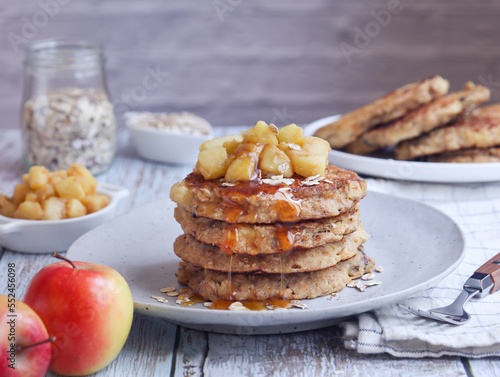 A plate of healthy rolled oat and apple pancakes topped with cooked diced apple pieces with sweet syrup. Its fruit and fork in scene with a bottle of oat grains in background 