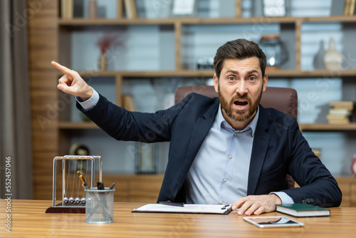 Принти на полотні Angry boss shouts at the camera to the employee points his finger to the side dismisses the subordinate, a businessman in a business suit sits at a table in a modern office