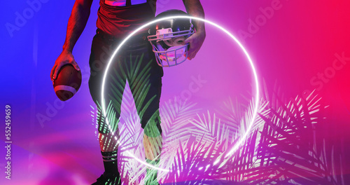Low section of american football player holding ball and helmet by illuminated circle and plants