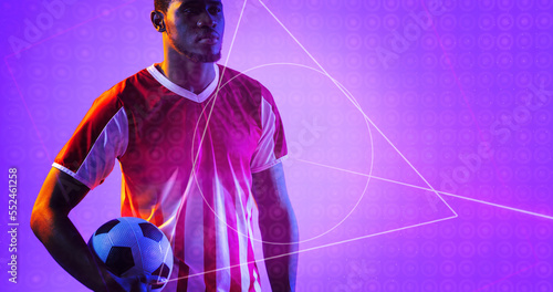 African american soccer player holding ball by geometric neon design over purple background