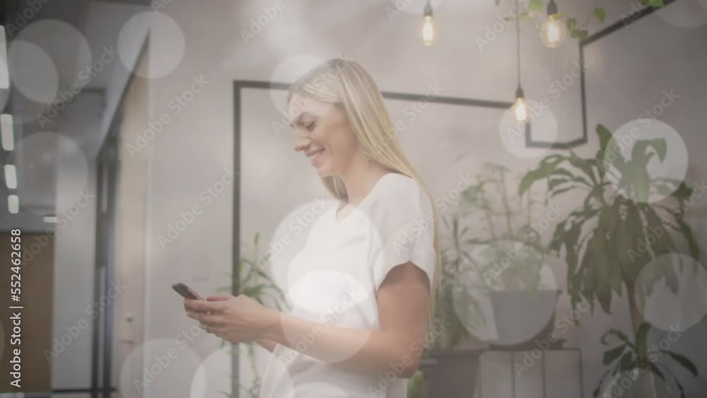 Animation of bokeh light spots over happy caucasian woman using smartphone in office Animation of bokeh light spots over happy caucasian woman using smartphone in office
