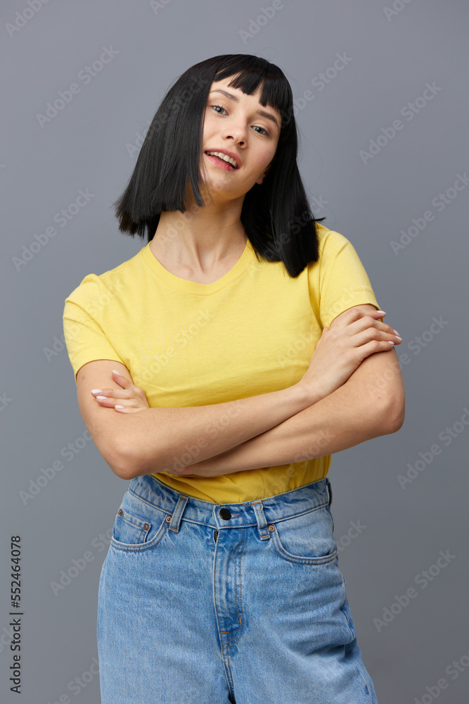 beautiful, attractive woman is standing in a yellow T-shirt and light jeans on a gray background with her arms crossed over her chest and smiling prettily at the camera. Vertical Studio Photography beautiful, attractive woman is standing in a yellow T-shirt and light jeans on a gray background with her arms crossed over her chest and smiling prettily at the camera. Vertical Studio Photography