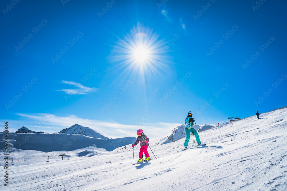 Mother teaching daughter how to ski, skiing down on a ski slope. Sunny day on ski winter holidays in Andorra, El Tarter, Pyrenees Mountains