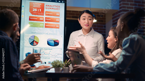 Asian business coach explaining statistics graphs visualization, giving presentation in meeting, holding clipboard. Diverse employees discussing marketing research analytics