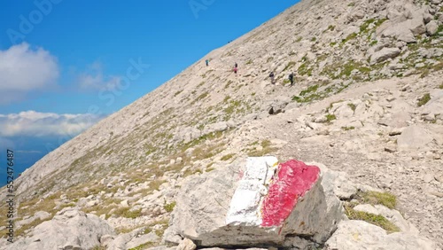 Marked direction signs painter on big stone in mountains peak, hiking important signs of red and white colour guiding people in right direction, blurred walking tourists on background. Special hiking photo