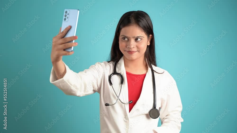 Portrait of a young female doctor taking selfie with mobile phone, Asian Indian woman doctor in apron isolated over blue studio background, taking photo with smartphone Portrait of a young female doctor taking selfie with mobile phone, Asian Indian woman doctor in apron isolated over blue studio background, taking photo with smartphone