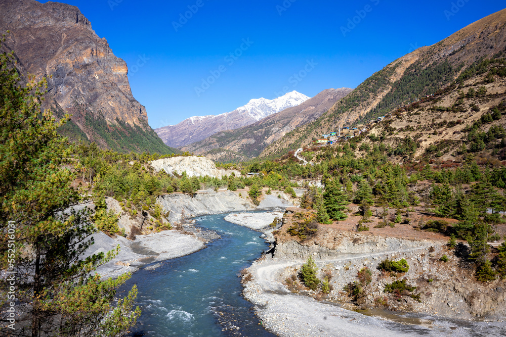 View of the valley in the mountains of Annapurna with a blue sky. A blue river flows through the valley and we see green trees on the slopes.