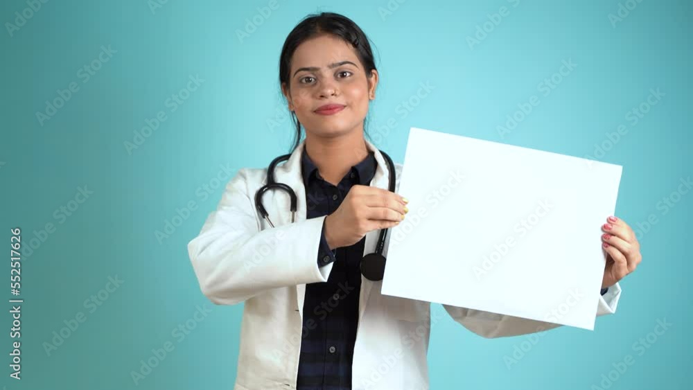 Portrait of a happy young female doctor holding blank white board, Cheerful Asian Indian woman doctor in apron and stethoscope showing empty white paper isolated over blue studio background Portrait of a happy young female doctor holding blank white board, Cheerful Asian Indian woman doctor in apron and stethoscope showing empty white paper isolated over blue studio background