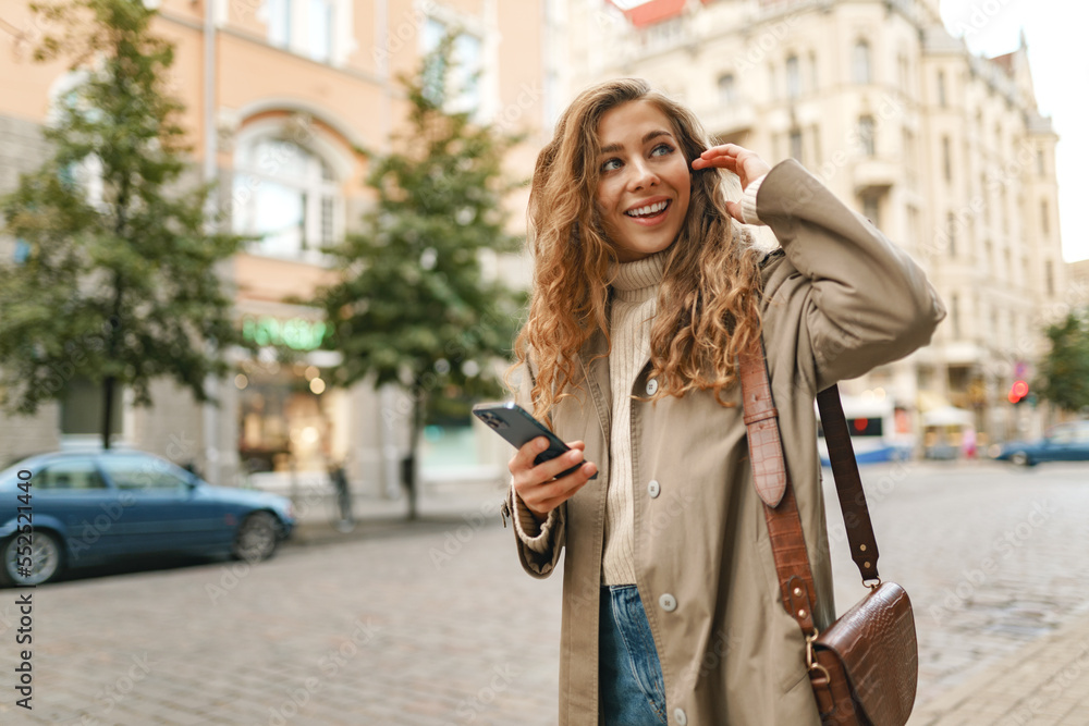 Smiling curly woman wearing warn coat walking down the street and using her phone