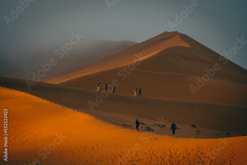 Touristen wandern über einen von der Morgensonne erleuchteten Grat in Richtung des nebelverhangenen Gipfels der Düne Big Daddy im Sossusvlei (Namibia)