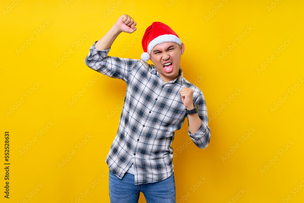 Smiling young Asian man wearing a plaid shirt in a Christmas hat posing doing winner gesture clenching fists on yellow background. celebration Christmas holiday and New Year concept Smiling young Asian man wearing a plaid shirt in a Christmas hat posing doing winner gesture clenching fists on yellow background. celebration Christmas holiday and New Year concept