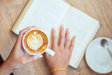 Top view of woman hand holding cup of latte while reading in cafe.