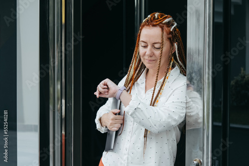 Woman with dreadlocks and laptop leaning against front door looking at her watch