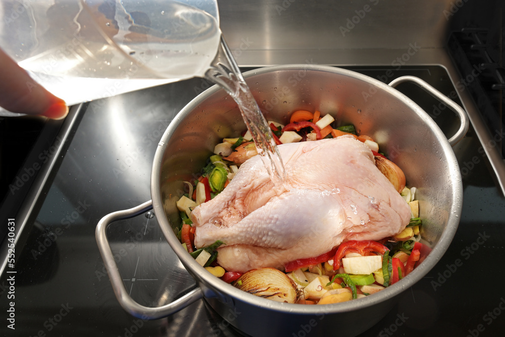 Whole raw chicken in a pot with chopped vegetables is poured with cold water for cooking a healthy soup against cold and flu, selected focus Whole raw chicken in a pot with chopped vegetables is poured with cold water for cooking a healthy soup against cold and flu, selected focus