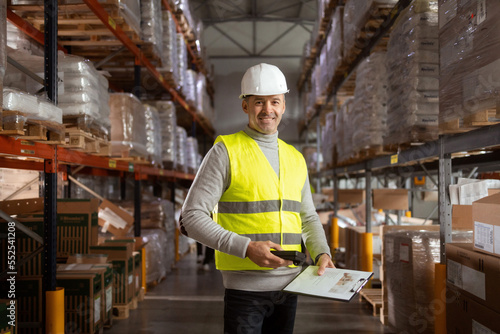 Portrait of a man working in warehouses. He is wearing protective workwear and holding a clipboard and barcode reader.