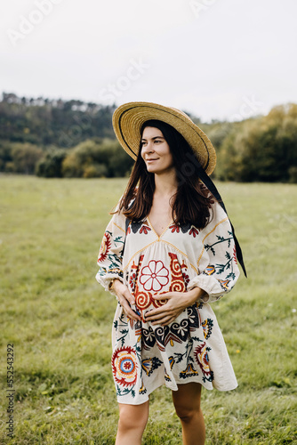 Pregnant woman outdoors, in nature, wearing a straw hat and a vintage flower embroidered dress, holding hands on belly.