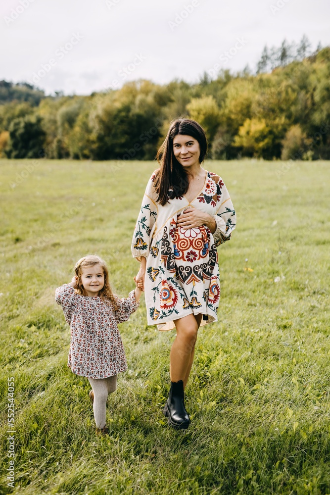 Obraz premium Pregnant woman walking with her toddler daughter in an open field, spending time together. Big sister concept.