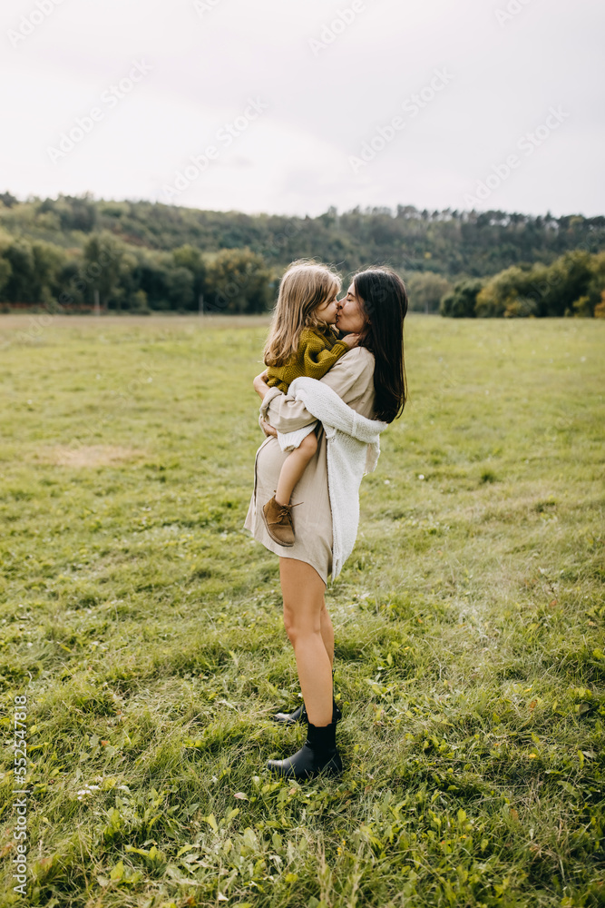 Pregnant woman and her toddler daughter spending time outdoors. Mother holding little girl in arms, touching noses. Pregnant woman and her toddler daughter spending time outdoors. Mother holding little girl in arms, touching noses.