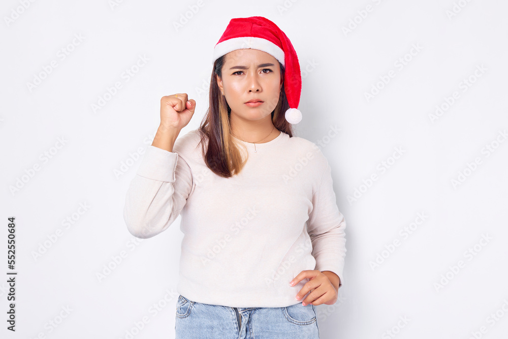 Attractive young Asian woman in Santa Claus hat clenching her hands first an angry expression on camera isolated over white background Attractive young Asian woman in Santa Claus hat clenching her hands first an angry expression on camera isolated over white background