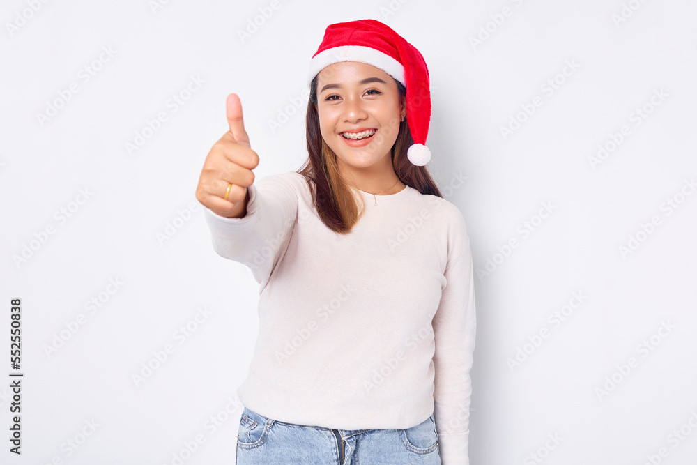 Portrait of smiling young Asian woman in Santa Claus hat showing thumbs up isolated over white background. celebration Christmas holiday and New Year concept Portrait of smiling young Asian woman in Santa Claus hat showing thumbs up isolated over white background. celebration Christmas holiday and New Year concept