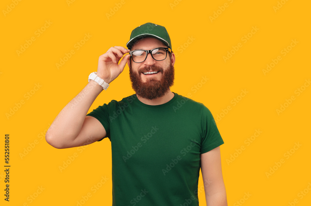 Fototapeta premium Portrait of a smiling delivery man wearing green uniform puts on some glasses over yellow background.