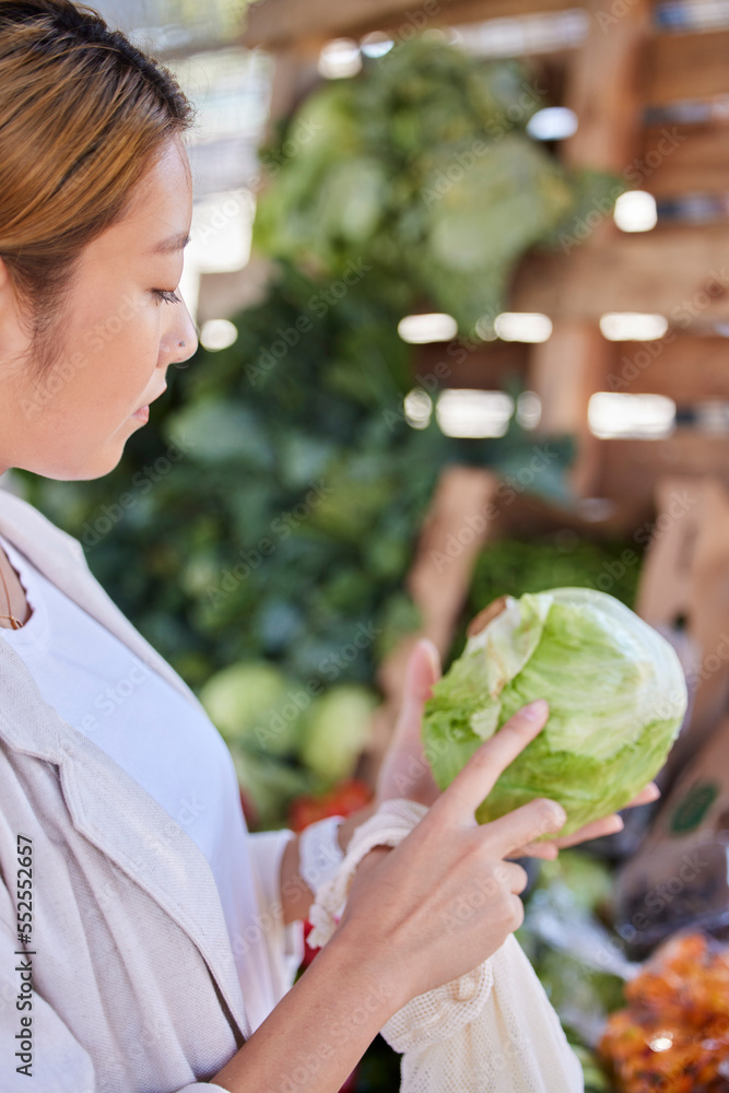 Obraz premium Grocery store, lettuce and Asian woman shopping and looking at vegetable quality and sale. Customer holding and checking vegetables price on salad promotion in a health food shop or supermarket