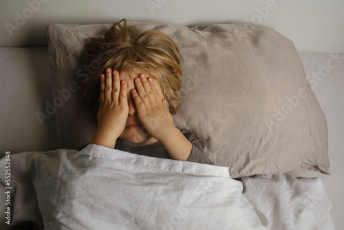 The child was scared before going to bed. Night terrors in a child. The kid covers his face with his hands in a fear. Children's experiences. Boy in the bed. View from above. Sad psychological state.