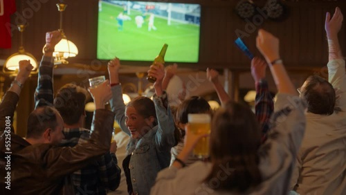 Group of Soccer Fans Cheering, Screaming, Raising Hands and Jumping During a Football Game Live Broadcast in a Sports Pub. Player in Blue Shirt Scores a Goal and Friends Celebrate. Slow Motion Footage photo