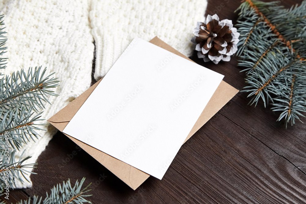 Obraz premium Christmas greeting card mockup with envelope, knitted sweater, fir tree branches and cone on brown wooden background. Blank New Year holiday card mock up with winter decoration