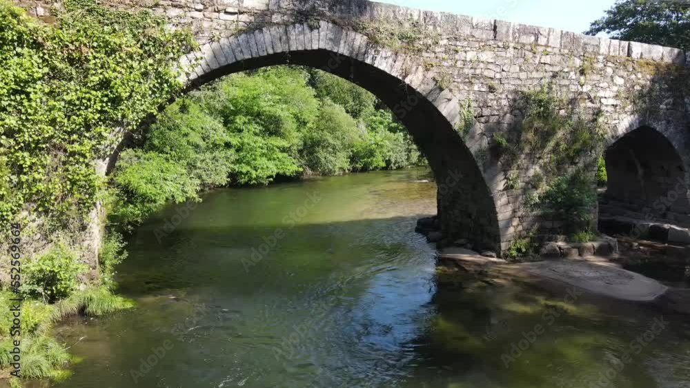 Drone video flying under a historic Roman bridge following the river in Galicia, Spain. Medieval old stone bridge covered with vegetation in the middle of the forest. Drone video flying under a historic Roman bridge following the river in Galicia, Spain. Medieval old stone bridge covered with vegetation in the middle of the forest.