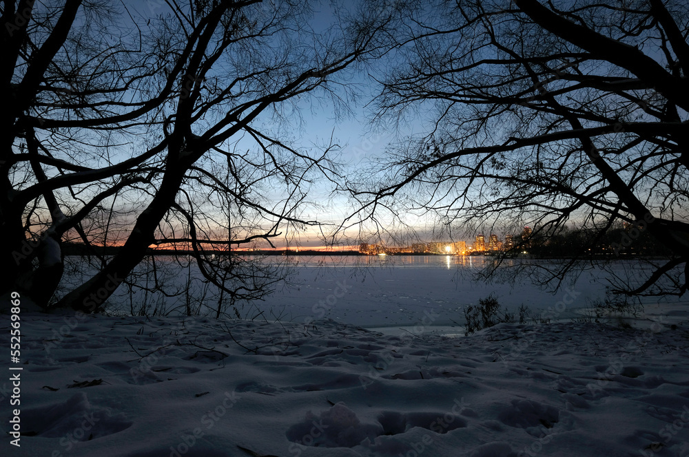 Beautiful night landscape after sunset with the bay in ice and urban residential houses with lighting after it, view through the tree branches silhouettes Beautiful night landscape after sunset with the bay in ice and urban residential houses with lighting after it, view through the tree branches silhouettes