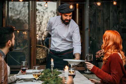 A male chef in uniform is serving a private elegant dinner to a group of people, People are smiling and welcoming the chef who is serving and bringing delicious food.