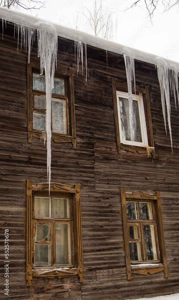 Growing, frozen icicles hang on the edge of the roof, winter or spring. Log wall of an old wooden house with windows. Large cascades of icicles in smooth, beautiful rows. Cloudy winter day, soft light