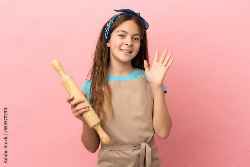 Little caucasian girl holding a rolling pin isolated on pink background saluting with hand with happy expression Little caucasian girl holding a rolling pin isolated on pink background saluting with hand with happy expression
