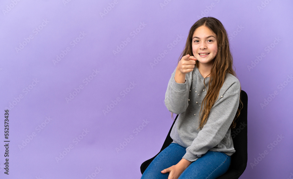 Little caucasian girl sitting on a chair isolated on purple background surprised and pointing front Little caucasian girl sitting on a chair isolated on purple background surprised and pointing front