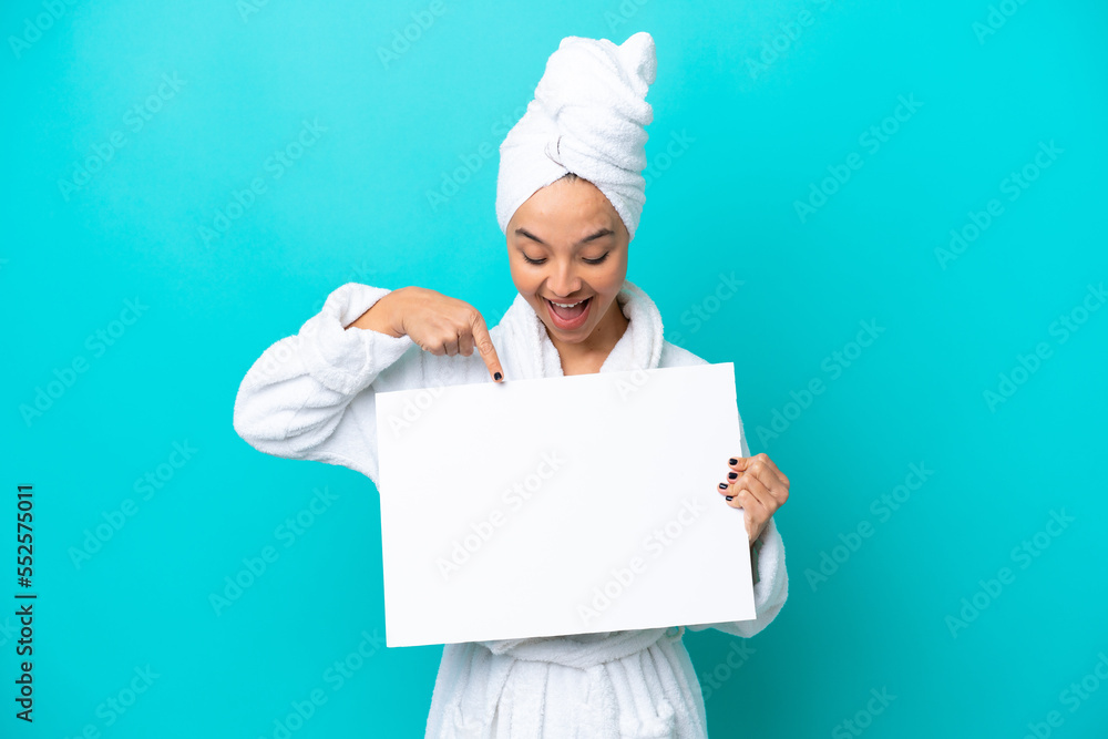 Young woman in a bathrobe with towel isolated on blue background holding an empty placard with happy expression and pointing it Young woman in a bathrobe with towel isolated on blue background holding an empty placard with happy expression and pointing it