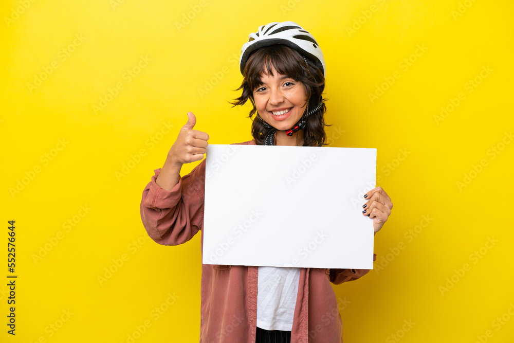 Young cyclist latin woman isolated on yellow background holding an empty placard with thumb up Young cyclist latin woman isolated on yellow background holding an empty placard with thumb up