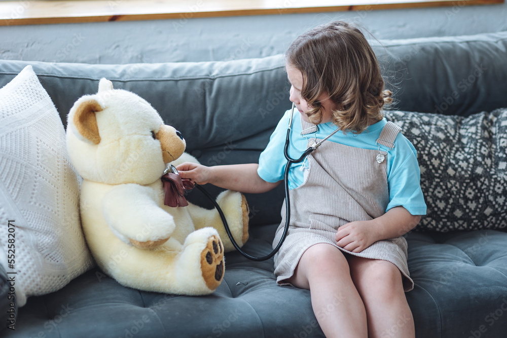 Cute little girl playing funny game being a doctor with her big fluffy teddy bear at home sitting on the sofa, checking heart beat with stethoscope. Cozy light Cute little girl playing funny game being a doctor with her big fluffy teddy bear at home sitting on the sofa, checking heart beat with stethoscope. Cozy light