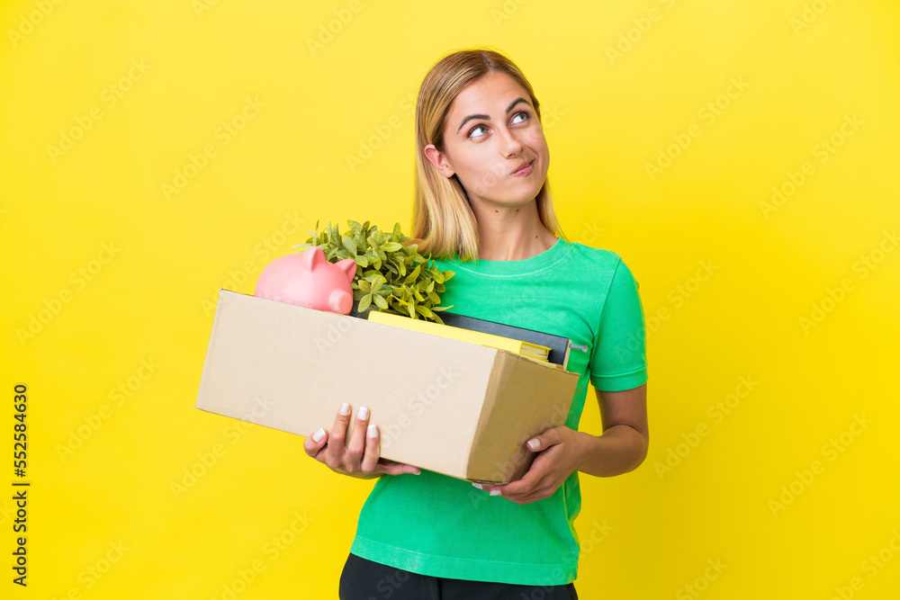 Young Uruguayan girl making a move while picking up a box full of things isolated on yellow background and looking up Young Uruguayan girl making a move while picking up a box full of things isolated on yellow background and looking up