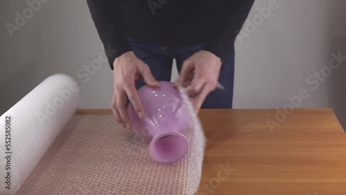 Close-up of a man's hands covering a glass vase with an air bubble wrap at a table in a warehouse