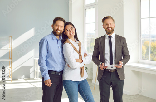 Young family decides to buy a house after a house tour. Portrait of a happy young couple together with a real estate agent standing in a big light room, looking at the camera and smiling