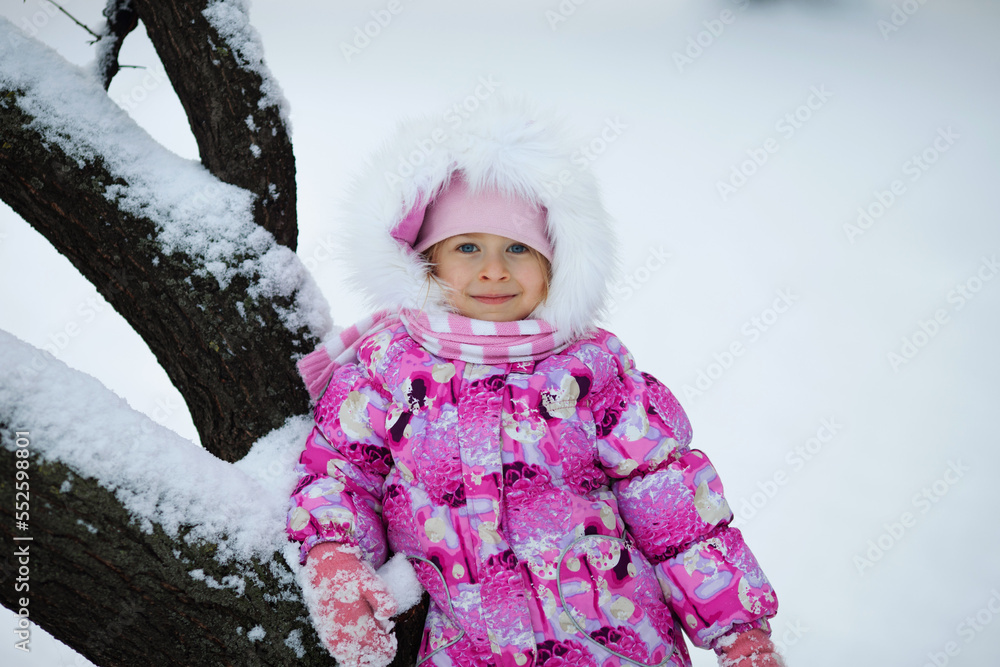 portrait of a little blue-eyed girl smiling happily. In bright winter clothes and a hood with fur on the background of a snowy park portrait of a little blue-eyed girl smiling happily. In bright winter clothes and a hood with fur on the background of a snowy park